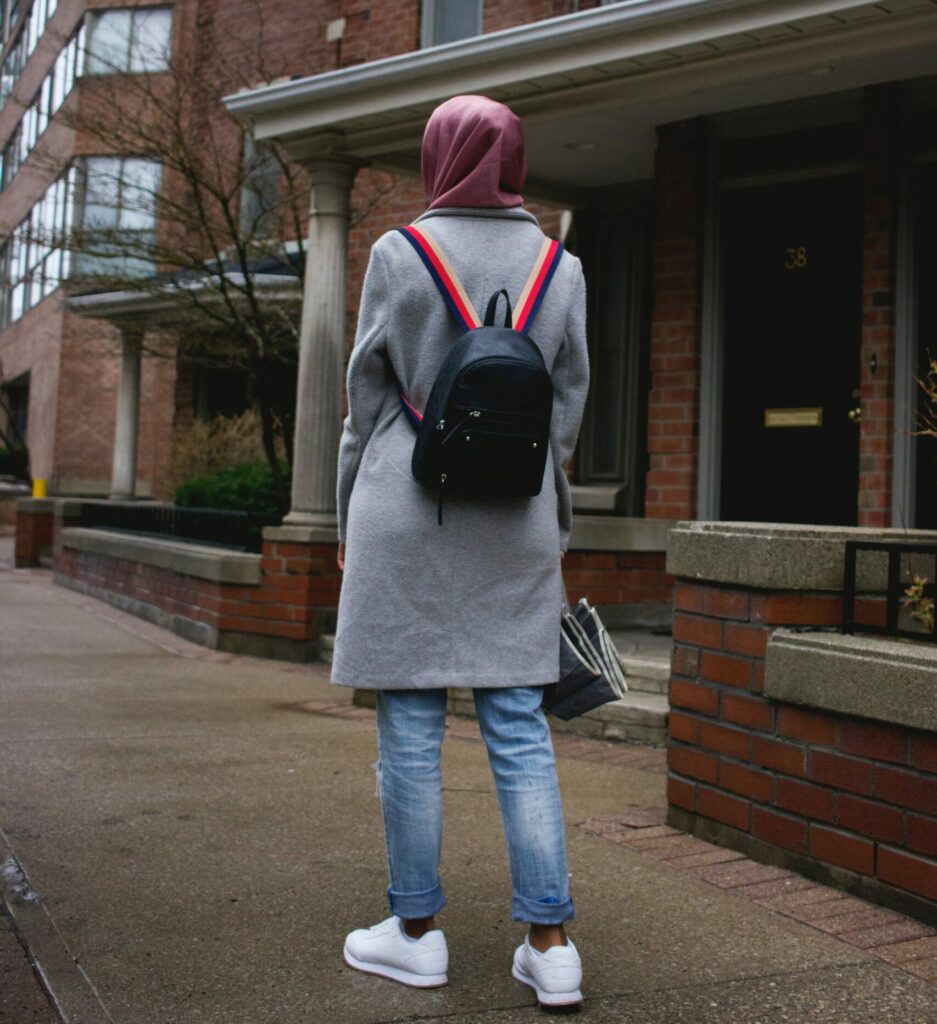 Mujer joven con pañuelo rosa en la cabeza y abrigo gris de pie en una acera frente a unos edificios de ladrillo. Está de espaldas a la cámara y lleva una pequeña mochila negra.