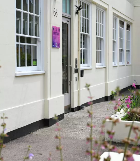 The ground floor of a white building with a grey door. Next to the door is a small square pink sign. Large sash windows have with mutin bars. In the foreground are green plants and pink flowers