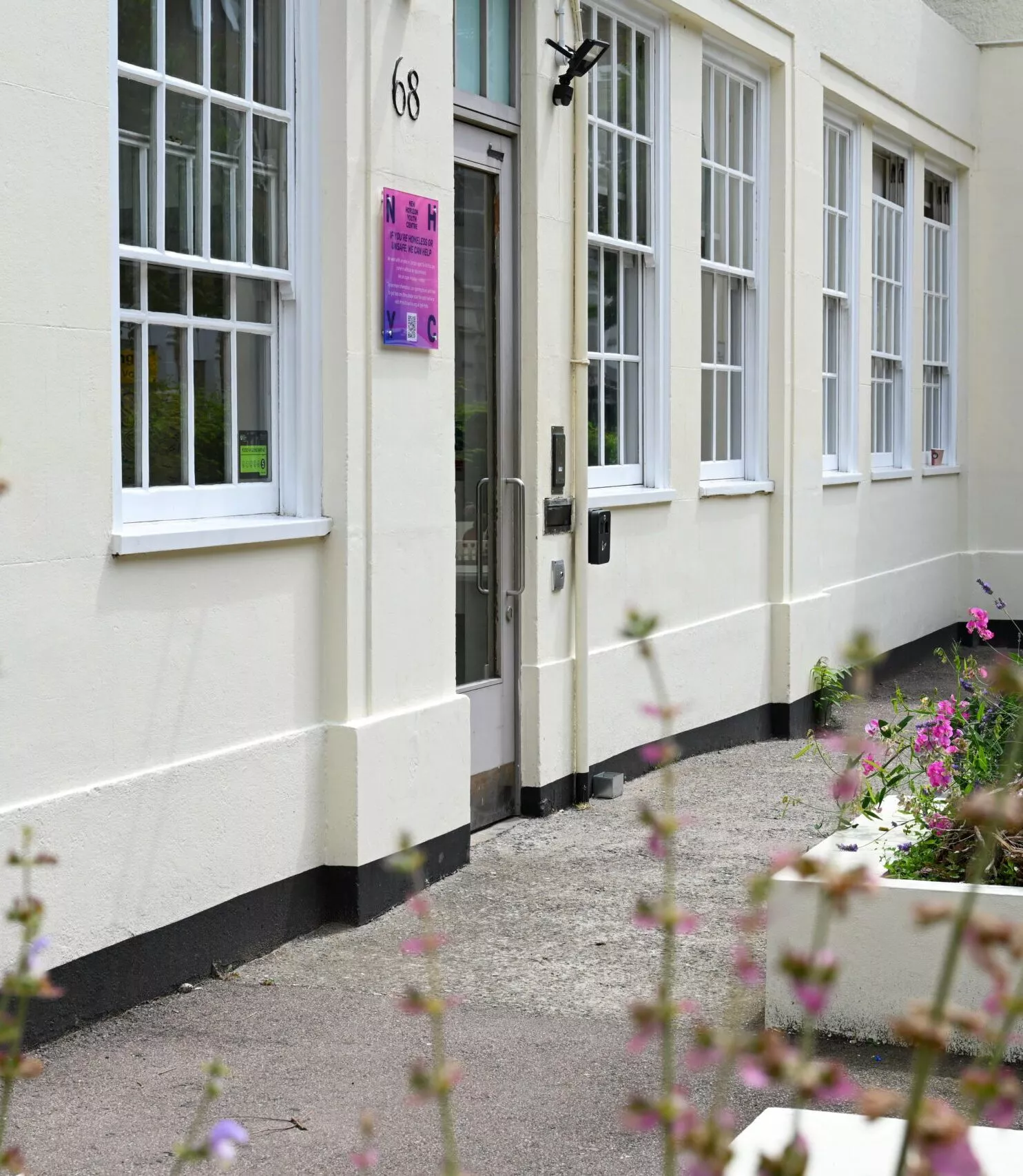 The ground floor of a white building with a grey door. Next to the door is a small square pink sign. Large sash windows have with mutin bars. In the foreground are green plants and pink flowers