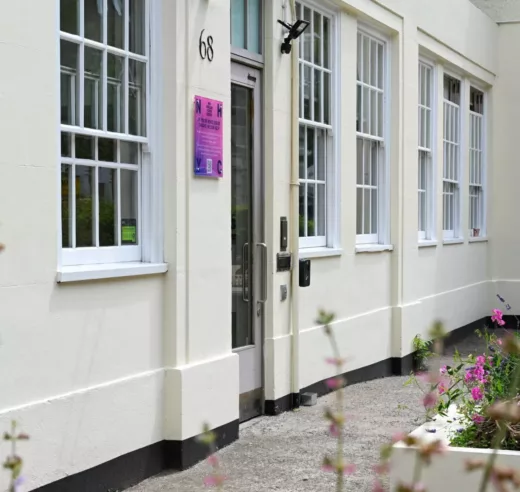 The ground floor of a white building with a grey door. Next to the door is a small square pink sign. Large sash windows have with mutin bars. In the foreground are green plants and pink flowers