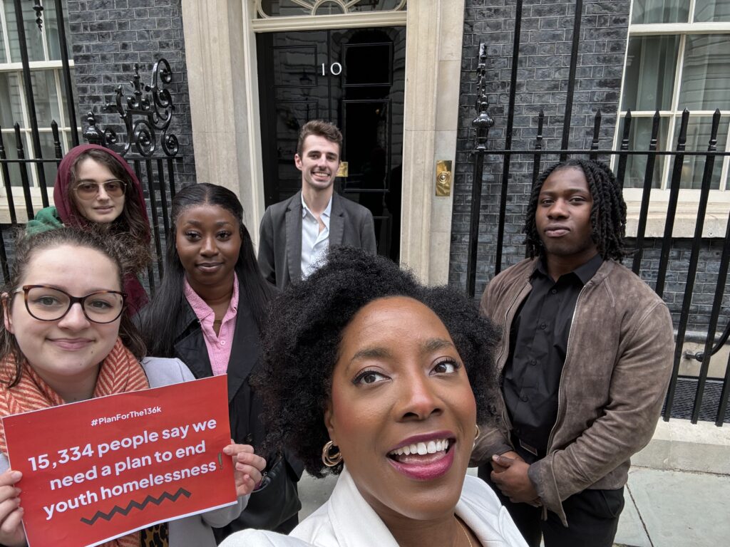 Selfie taken outside Number 10 Downing Street. The four young people are in the back, at the front is Lily smiling wearing a white coat and Polly holding a red sign saying "15,334 people say we need a plan to end youth homelessness!"
