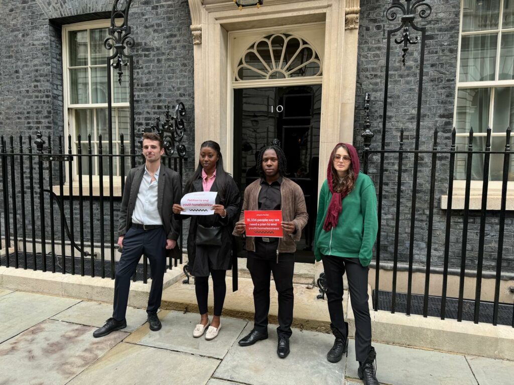 Four young people stand in front of Number 10 Downing Street door. Felix is wearing a grey suit jacket and pale shirt; Deborah a black coat and a pink shirt; Andrew is wearing a black shirt, trousers and brown jacket and Stella wears a green jumper and a dark red headscarf.