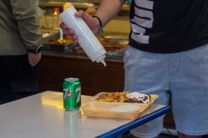 Man squirting mayonnaise on burger and chips in a takeaway container. You can only see the man's blue shorts and part of his black t-shirt.