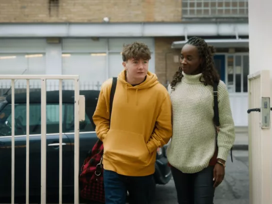 a young man with a yellow hoodie being led through a gate by a woman with a white jumper