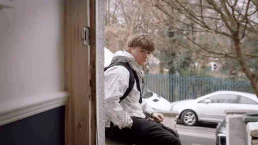 Young man wearing a white cagoule sitting outside someone's front door as it is opened from the inside.