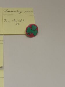 A pink and green badge with a shamrock on it attached to a post it note with Faraday's Law equation on it, against a plain white background