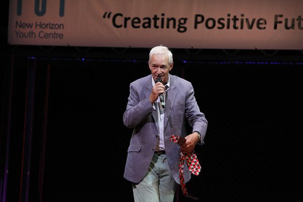 Jon Snow, a white man with white hair and a grey jacket is on stage with a microphone. In his hand he holds a red tie.