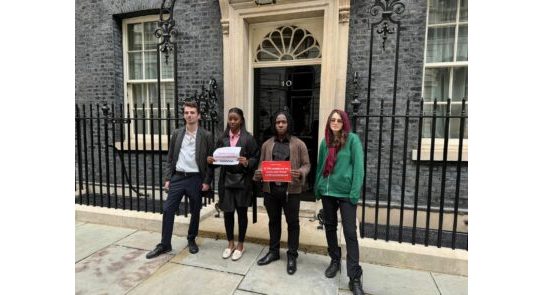 Photo of 4 young people outside Number 10 Downing Street. They are holding red and white pieces of paper to represent a petition hand in