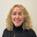 Photograph of Alice Godfrey, a white woman with shoulder length curly hair wearing a black roll neck jumper