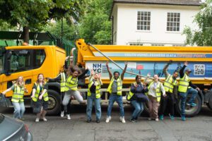 Photo of a big yellow McGee tipper truck with NHYC branding on the side and 9 people in front of it jumping and laughing wearing high vis jackets