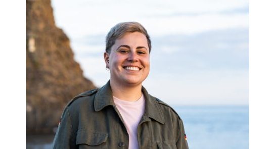 Young adult gender neutral with short hair - joyful smile against a seaside backdrop - serene outdoor moment captured at dusk.