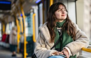 Young white woman looks out of a bus window wearing a green scarf and beige jacket. She has long brown hair with a fringe