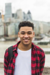 A young mixed race man is smiling in front of the Thames with City of London buildings in the background. He is wearing a bight red and black checked shirt over a white t-shirt, has a back pack slung over one shoulder and short brown hair.